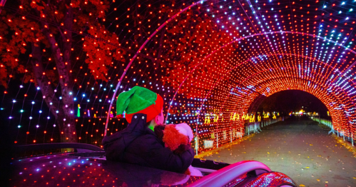A child in a festive hat gazes at a colorful light tunnel from a car roof, surrounded by vibrant holiday decorations.