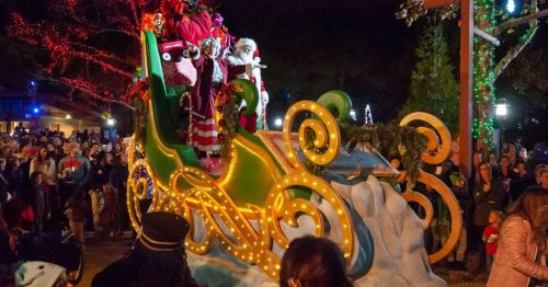 A festive parade scene featuring Santa Claus on a brightly lit sleigh, surrounded by a cheering crowd at night.
