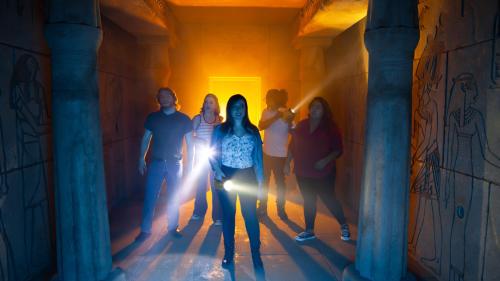 A group of six people exploring a dimly lit, ancient-looking corridor with flashlights, surrounded by hieroglyphs.