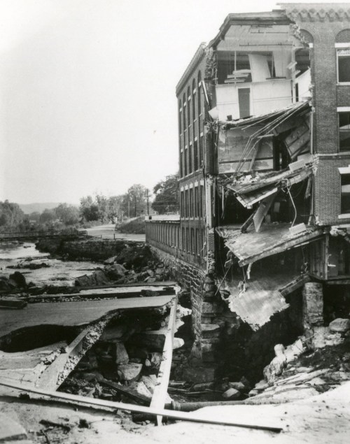 Black and white photo of a partially collapsed building near a river, showing significant structural damage.