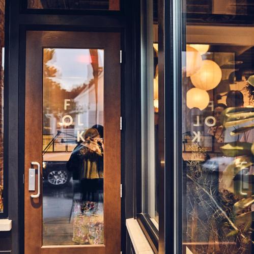 A wooden door with "FOLK" etched on it, reflecting a person and warm interior lights.