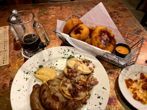 A plate of sausage, potatoes, and sour cream, with a basket of onion rings and a glass of dark beer on a table.