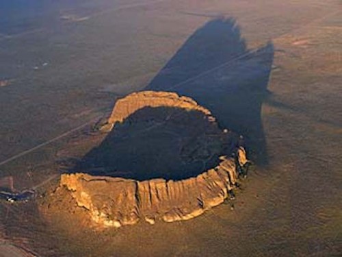 Aerial view of a large, flat-topped rock formation casting a long shadow over the surrounding landscape.