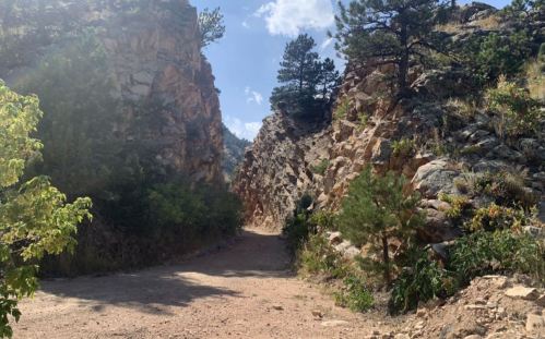 A narrow dirt path winds through rocky cliffs, surrounded by trees and greenery under a bright blue sky.