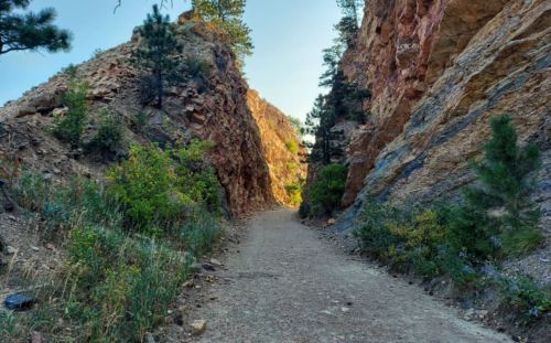 A narrow dirt path winds between steep, rocky cliffs, surrounded by greenery and trees under a clear blue sky.