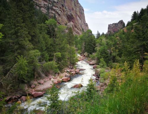 A serene river flows through a lush green valley, surrounded by rocky cliffs and trees under a cloudy sky.