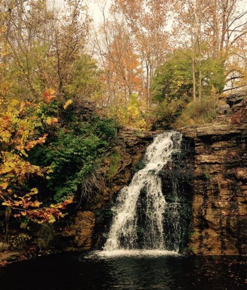 A serene waterfall cascades over rocky cliffs, surrounded by vibrant autumn foliage and lush greenery.