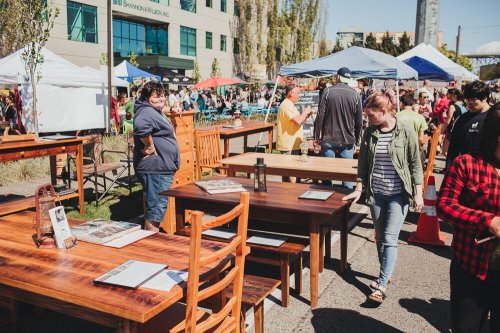 A bustling outdoor market with wooden furniture displays and people browsing under colorful tents.
