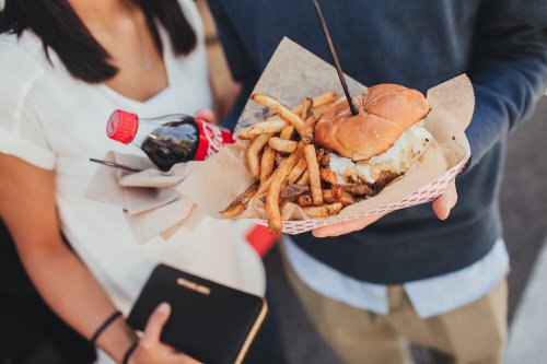 A person holds a tray with a burger, fries, and a bottle of soda, while another person stands nearby.