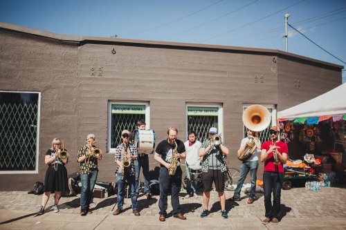 A lively brass band performs outdoors, with musicians playing various instruments in front of a colorful market.