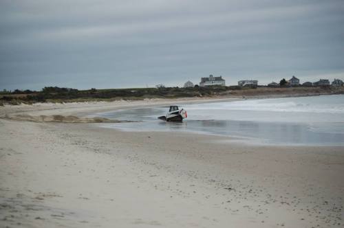 A small boat stranded on a sandy beach under a cloudy sky, with distant houses along the shoreline.