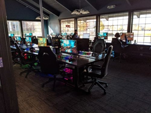 A gaming room with multiple desks, each featuring computers and colorful lighting, occupied by several players.