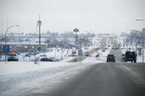 A snowy road stretches into the distance, lined with shops and vehicles under a gray sky.