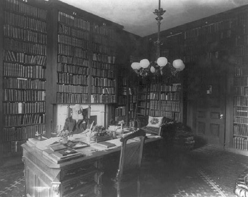 A vintage study with a large wooden desk, bookshelves filled with books, and a chandelier overhead.