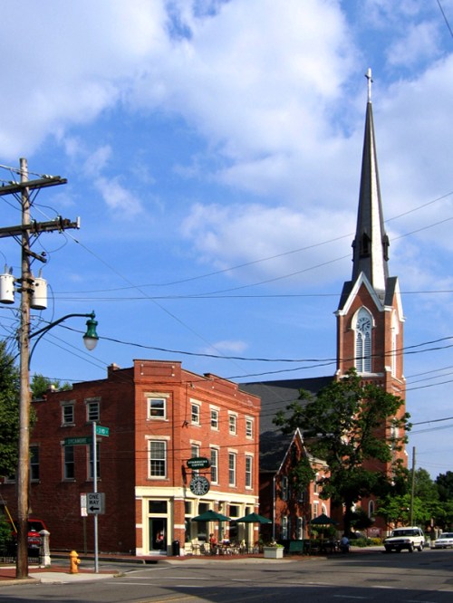 A historic brick church with a tall steeple stands next to a quaint building on a sunny day with blue skies.