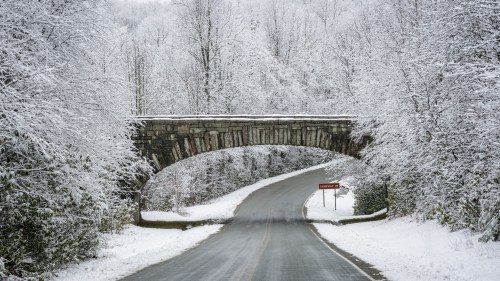 A snow-covered road curves under a stone bridge, surrounded by trees blanketed in white.