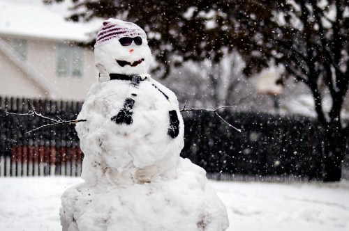 A cheerful snowman wearing sunglasses, a striped hat, and a scarf stands in falling snow.