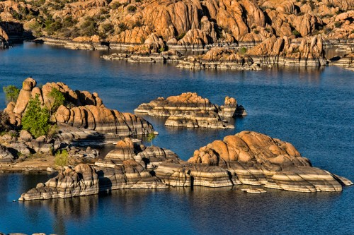 Rocky formations rise from a serene blue lake, surrounded by rugged terrain and clear skies.