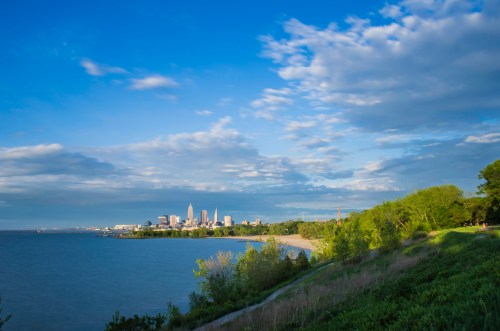 A scenic view of a city skyline by the water, with lush greenery and a blue sky filled with clouds.