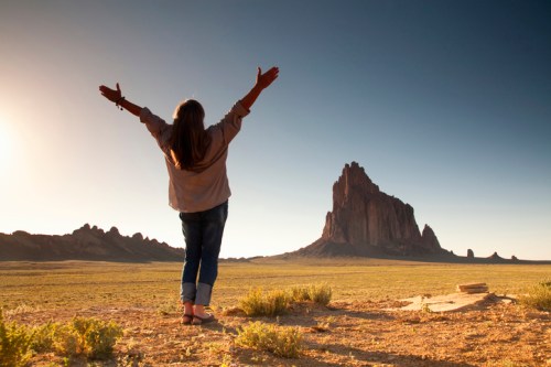 A person stands with arms outstretched in a vast landscape, facing a prominent rock formation under a clear sky.