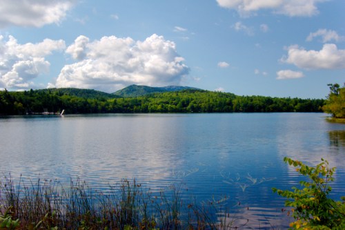 A serene lake surrounded by lush green hills and fluffy clouds in a blue sky, reflecting on the water's surface.