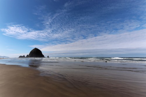 A serene beach scene with gentle waves, a rocky outcrop, and a blue sky with scattered clouds.