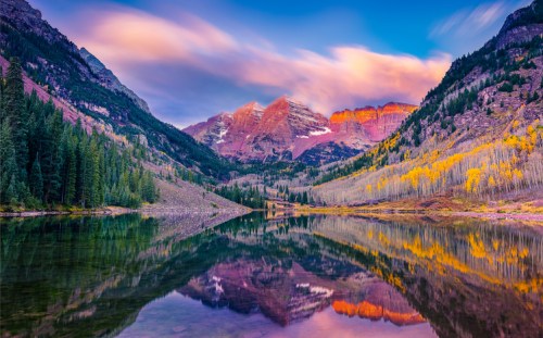 A serene mountain landscape at sunset, with colorful peaks reflected in a calm lake surrounded by trees.