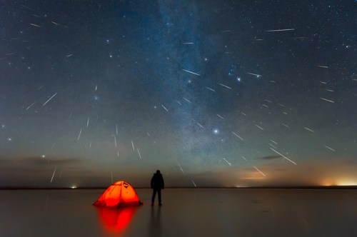 A person stands by a glowing orange tent under a starry sky filled with shooting stars and the Milky Way.