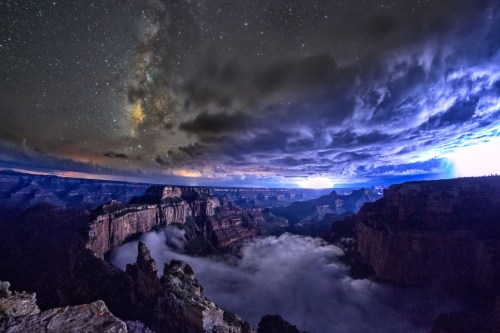 A stunning night sky over a canyon, with the Milky Way visible and clouds illuminated by distant lightning.