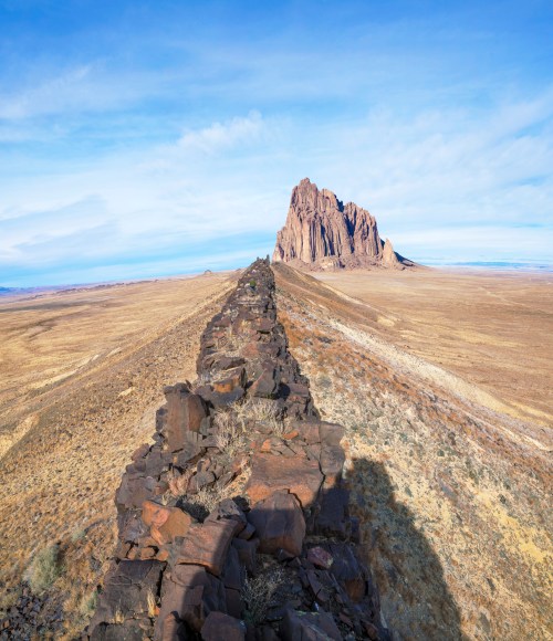 A rocky ridge leads to a towering rock formation under a blue sky, surrounded by vast, arid landscape.