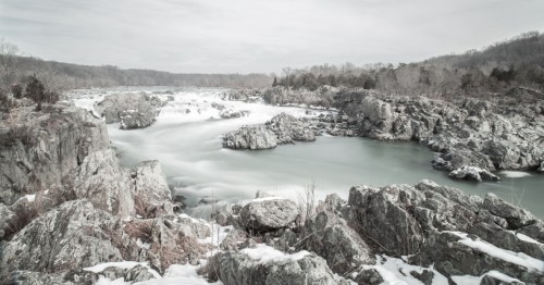 A serene winter landscape featuring a river flowing through rocky terrain, surrounded by snow-covered trees.