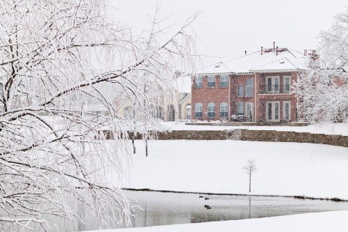 A snowy landscape featuring a brick house, a frozen pond, and bare trees, creating a serene winter scene.