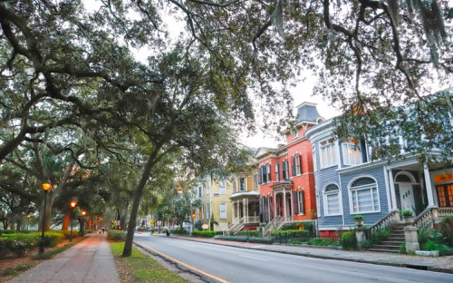 A tree-lined street with historic homes in various colors, illuminated by street lamps at dusk.
