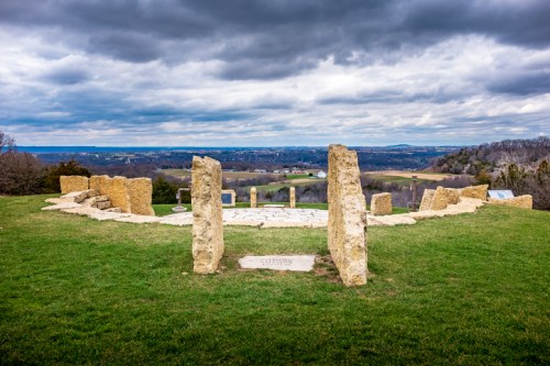 A stone circle on a grassy hilltop with a cloudy sky and distant landscape views.