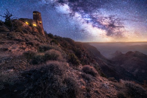 A starry night sky over a canyon, featuring a historic stone tower illuminated against the Milky Way backdrop.