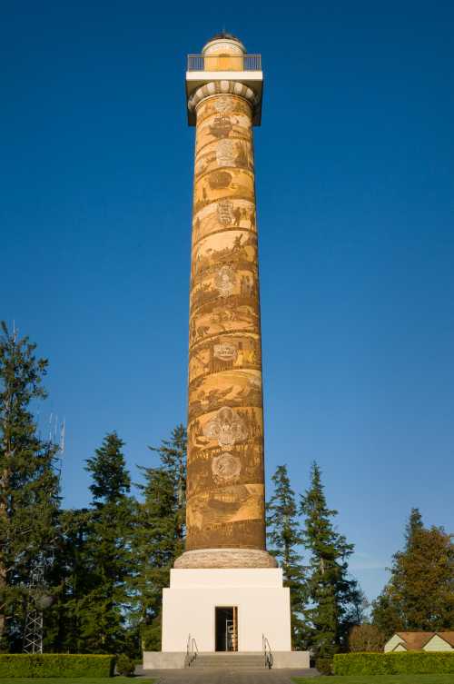 A tall, decorated column with murals stands against a clear blue sky, surrounded by trees.