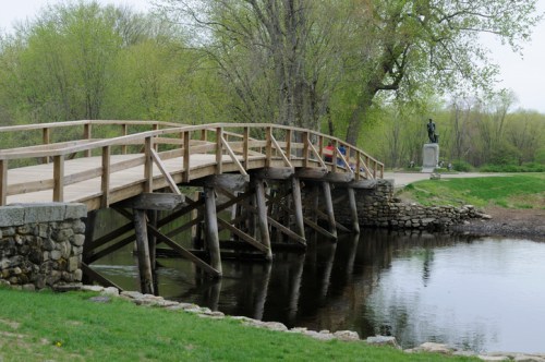 A wooden bridge over a calm river, surrounded by greenery, with a statue visible in the background.