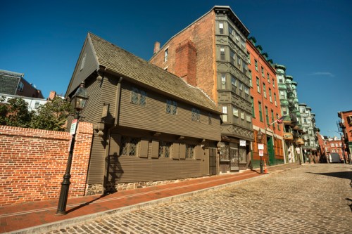 Historic building with a wooden facade next to modern brick and glass structures on a cobblestone street. Clear blue sky.