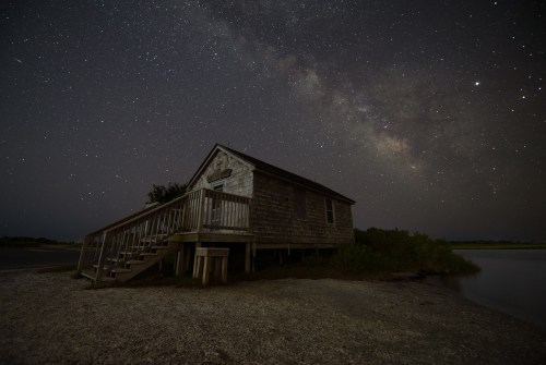 A rustic cabin by a calm waterway under a starry night sky, featuring the Milky Way in the background.