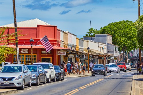 A bustling street scene with shops, pedestrians, and parked cars, featuring an American flag and clear blue skies.