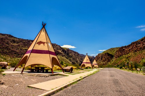 Three colorful tipis stand along a road, surrounded by rocky mountains and a clear blue sky.