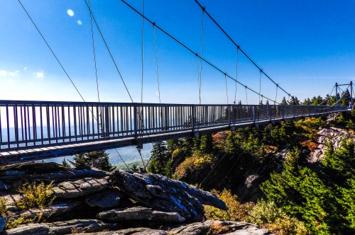 A suspension bridge spans a rocky landscape, surrounded by trees and a clear blue sky.