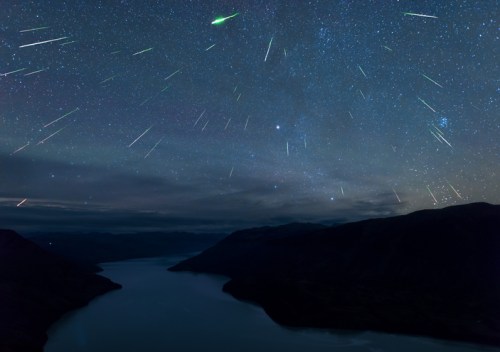 A starry night sky filled with shooting stars over a calm river and mountains in the background.