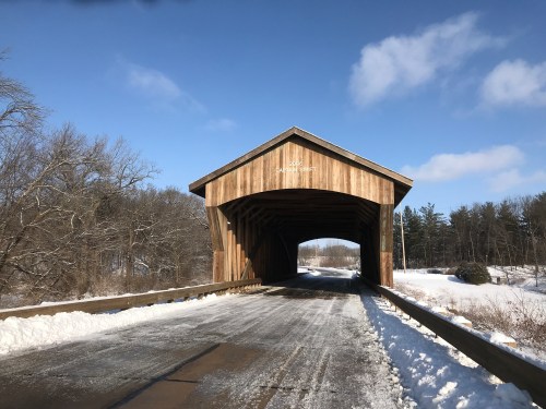 A snow-covered road leads into a wooden covered bridge under a clear blue sky. Trees line the background.