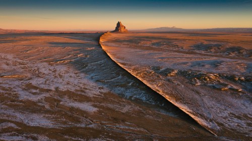 A dramatic landscape featuring a rocky formation rising from a vast, flat terrain under a colorful sky at sunset.