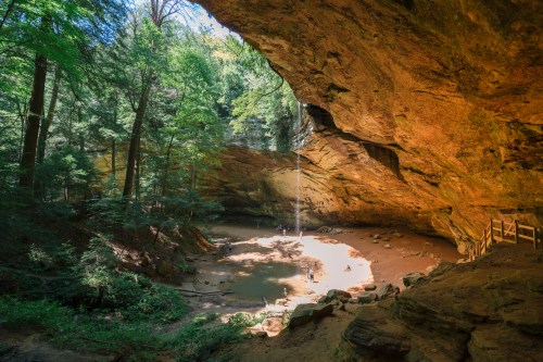 A serene cave with a waterfall, surrounded by lush green trees and a sandy area below, inviting exploration.