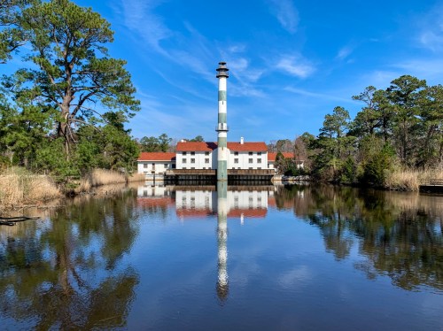 A lighthouse and building reflected in calm water, surrounded by trees under a clear blue sky.