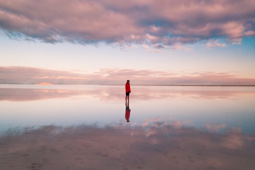 A person stands alone on a reflective salt flat at sunset, surrounded by colorful clouds and calm water.