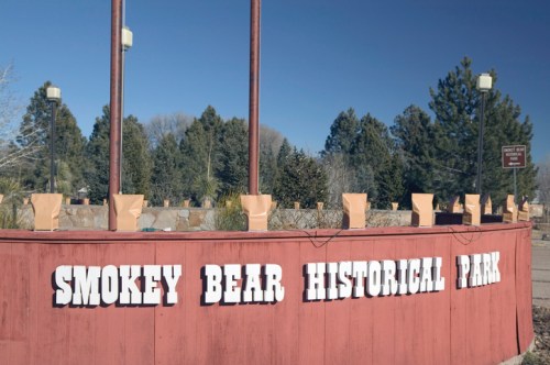 Sign for Smokey Bear Historical Park with brown paper bags on posts, surrounded by trees and clear blue sky.