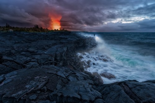 A dramatic coastline with crashing waves, dark volcanic rock, and a fiery orange glow from a distant eruption under stormy skies.